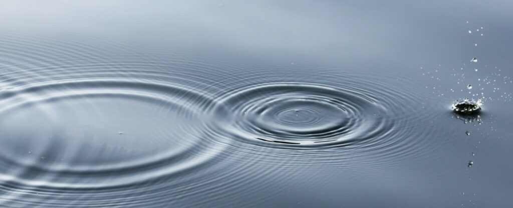 Ripples on a calm water surface, with a single droplet creating concentric circles and splashing nearby.