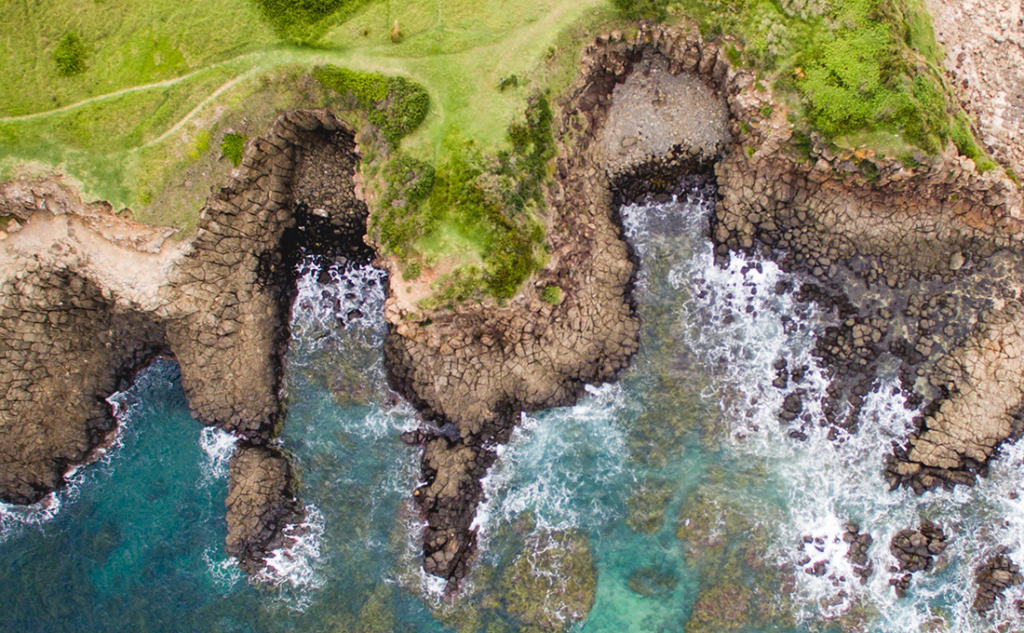Aerial view of rugged coastline with rocky formations and waves crashing against the shore, surrounded by lush green grass.