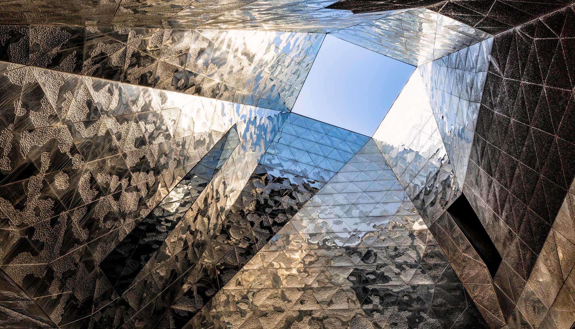 A geometric architectural interior with reflective surfaces converging towards a bright blue sky opening above.