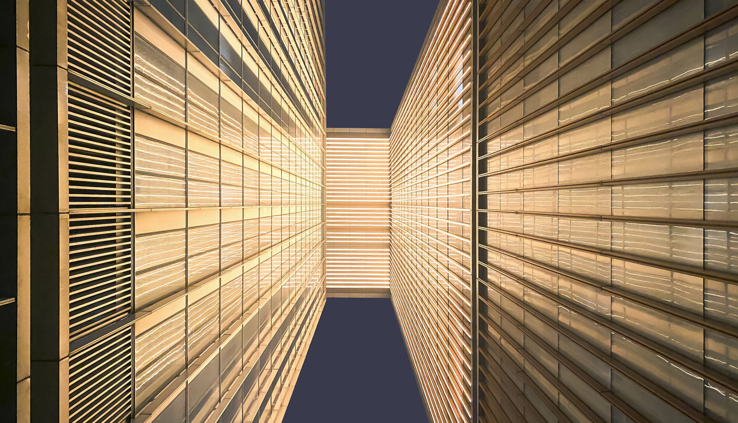 Looking up between two modern buildings, showcasing illuminated facades and sleek lines against a dark sky.