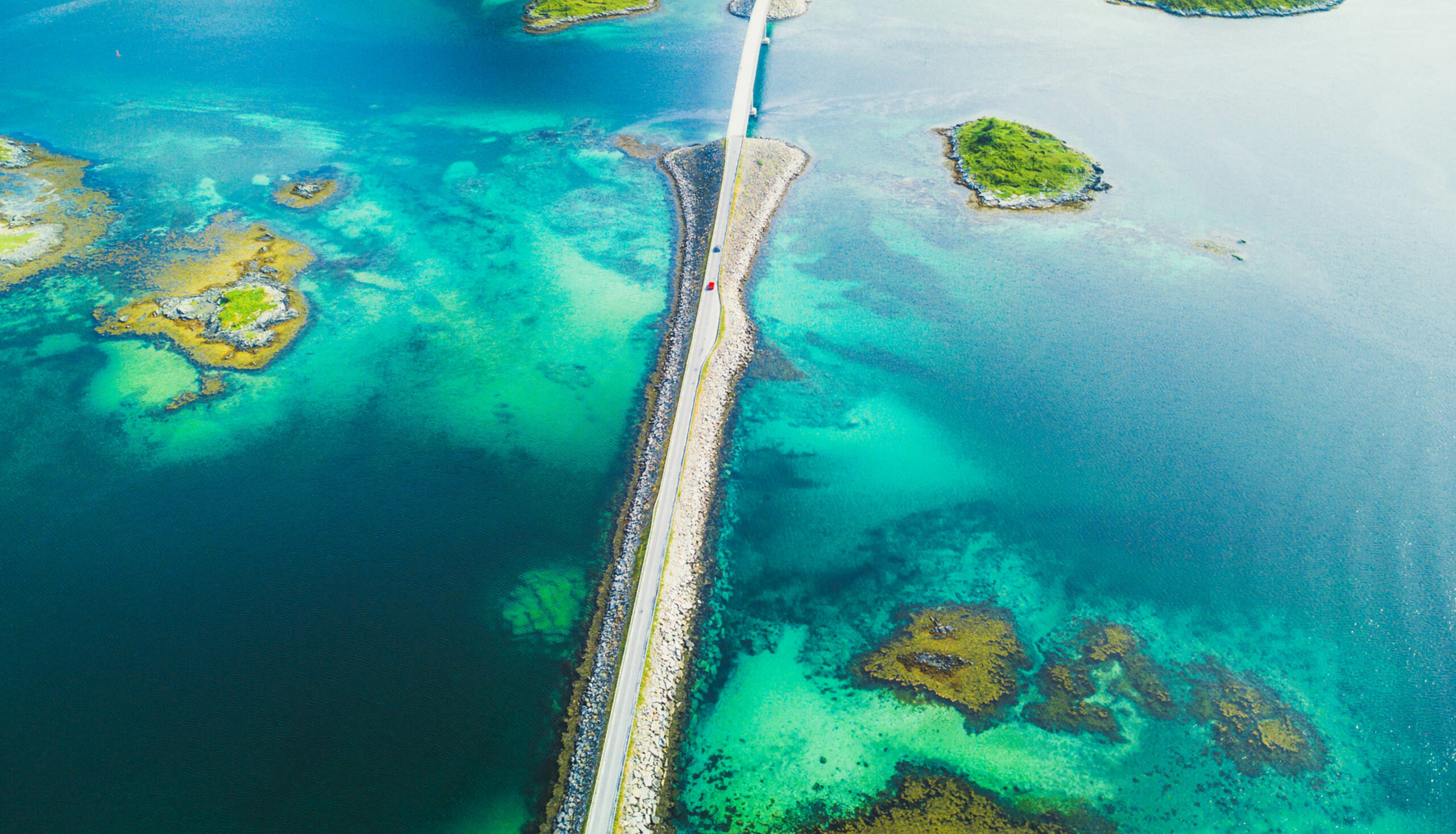 Ariel view of scenic road over turquoise waters, with lush islands and rocky shores surrounding it.