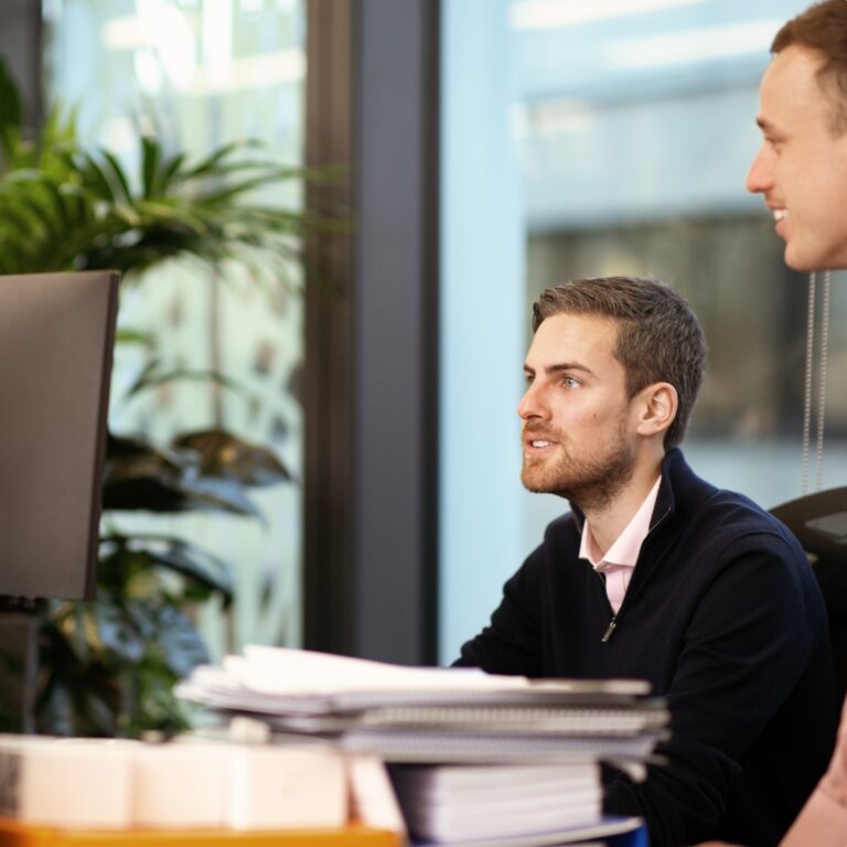 Male officer worker at his desk in conversation with another colleague.