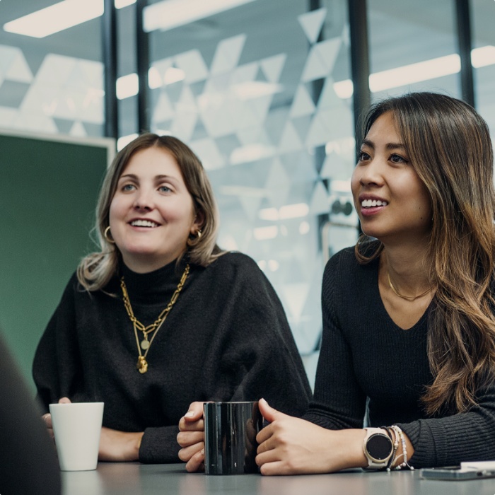 Two women having coffee at a table in an office, engaging in conversation.